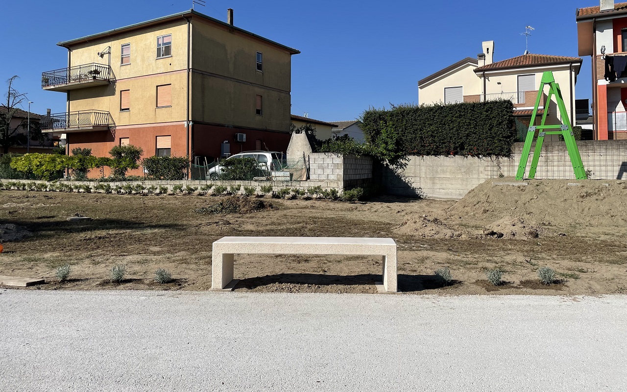 panca in cemento dalle linee pulite lungo il camminamento pedonale dall'interno del parco Vassallo della città di Rosolina, con nuova siepe di pittosporum tobira e rose rampicanti, e l'arredo ludico a teleferica